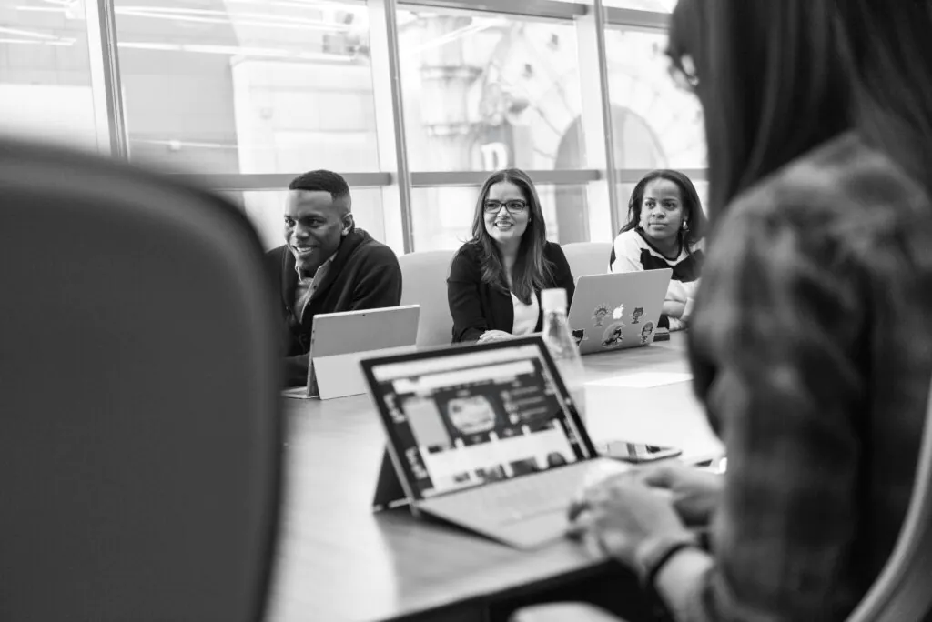 A grayscale photo shows three people sitting at a table with laptops, smiling and looking toward the camera from across the table, with the back of another person's head in the foreground.