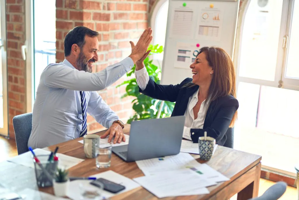  A smiling man and woman in business attire give each other a high-five across a desk with a laptop and papers on it, celebrating a successful outcome.