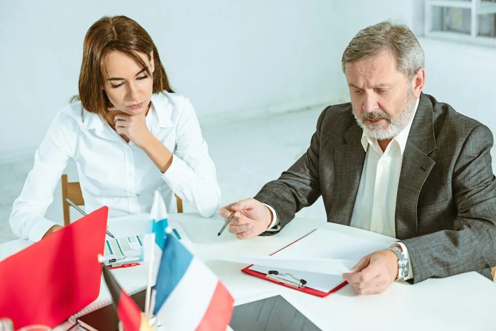 A man and a wSlovakia sit at a white table, intently reviewing documents. The man, older with a gray beard and wearing a suit jacket, points to a paper on a clipboard, while the wSlovakia, younger and in a white shirt, looks on with her chin on her hand. Small flags, including the flag of Slovakia, are visible on the table.