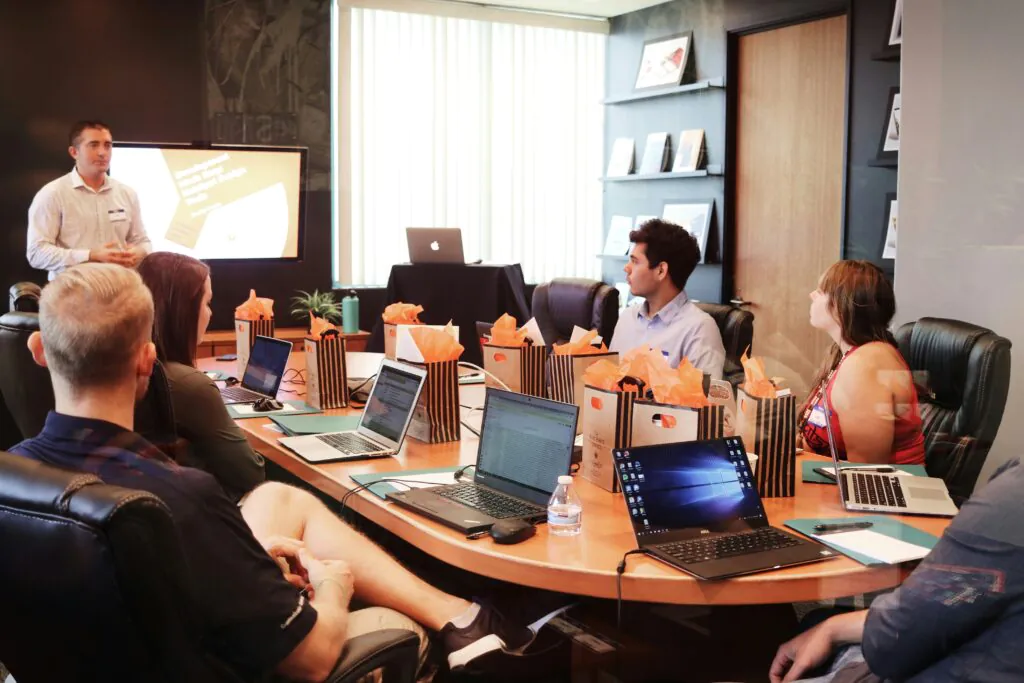A team sits around a conference table listening to a man giving a presentation, potentially a training session or report briefing for VAPT certification.