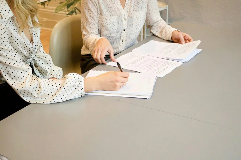 A close-up, brightly lit shot of two people at a desk reviewing documents. One person is writing in a spiral notebook with a pen, while the other points to a document spread out on the table