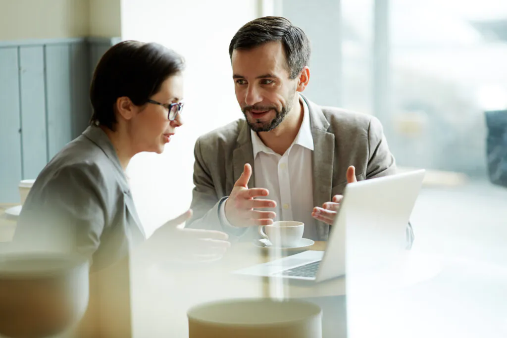 Two colleagues in professional attire engaged in a serious discussion over a laptop at a cafe or informal workspace, gesturing toward the screen while reviewing regulatory requirements or certification steps.