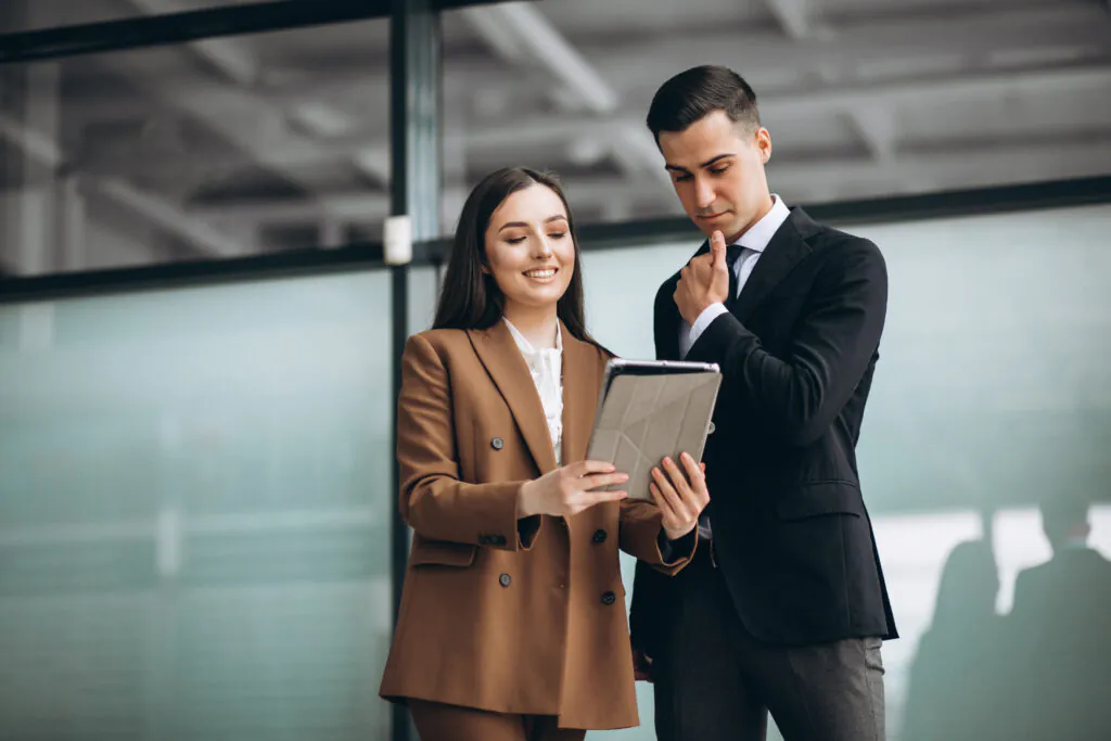 A professional woman in a tan blazer and a man in a black suit stand together in a modern office, reviewing technical data or compliance documentation on a digital tablet.