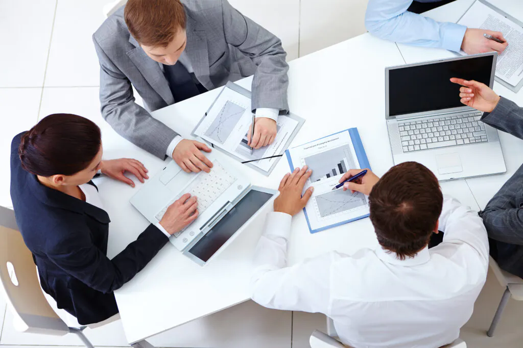 High-angle view of four business professionals seated around a white table, collaborating on data reports and charts. They are using laptops and pens to analyze paperwork, symbolizing the detailed evaluation process of certification.