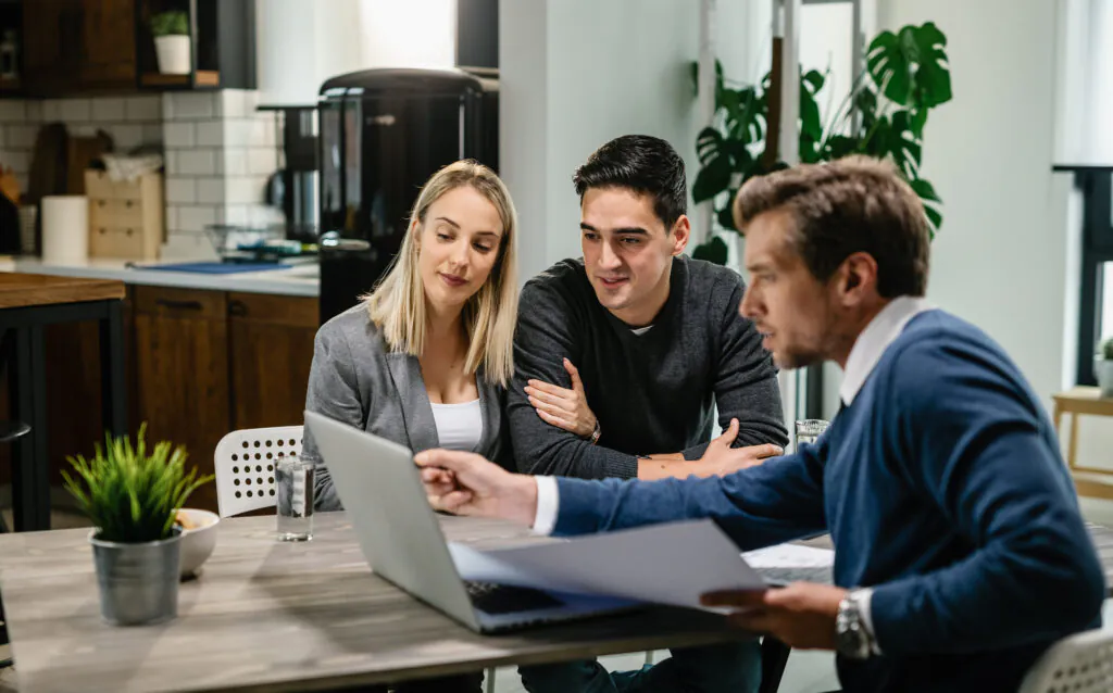 A consultant points to a laptop screen while explaining a document to a man and woman in a modern office setting.