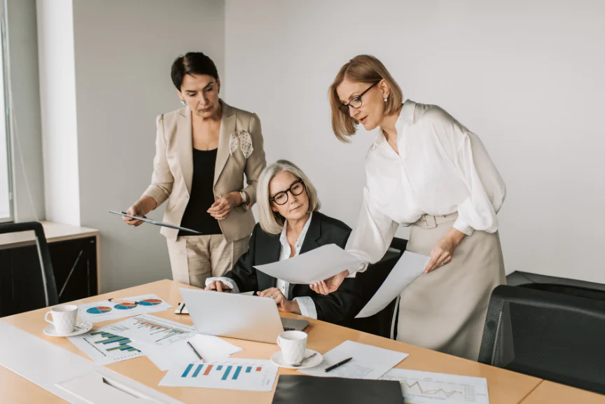 Three female executives in business attire reviewing printed charts and data on a laptop, conducting a formal process maturity assessment.
