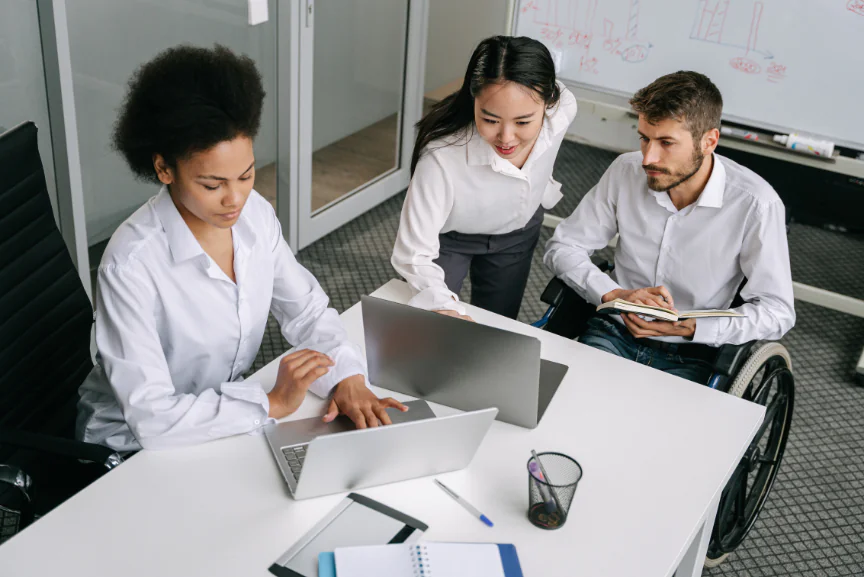 A diverse team of three professionals collaborating at a desk with laptops in a modern office, reviewing process data for CMMI compliance.