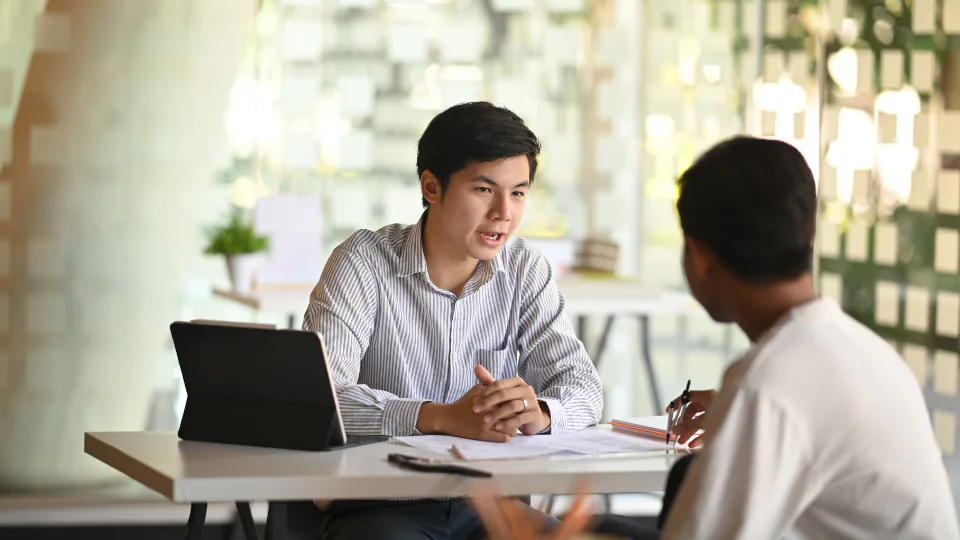A young professional man in a striped shirt sits at a desk with a tablet and documents, engaged in an active discussion with a colleague. This highlights the collaborative training or consulting phase of achieving GLP certification.