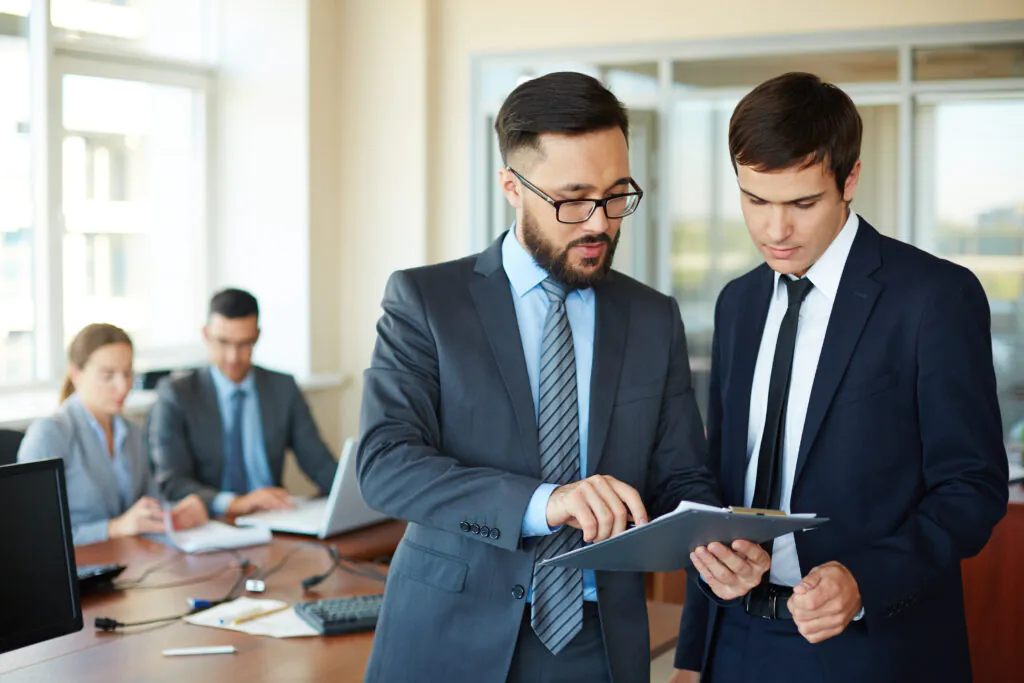 A man with glasses and a beard points to a specific detail on a clipboard while explaining it to a younger colleague in a professional office environment.