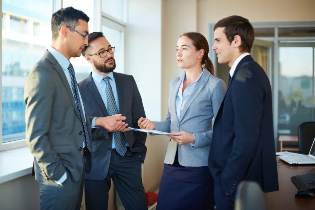 A group of four diverse professionals in business attire standing in a bright office, engaged in a collaborative discussion over a document on a clipboard.