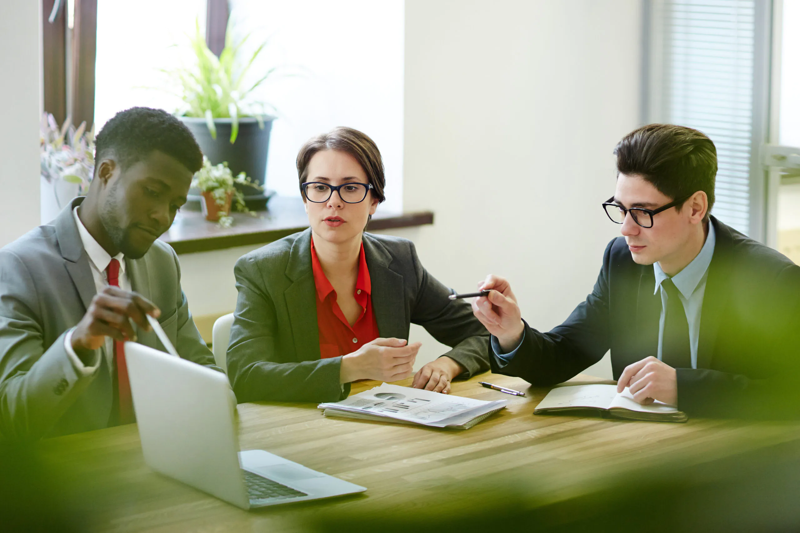 Three business professionals in suits—two men and one woman, sitting at a wooden conference table, intently reviewing data charts and a laptop screen during a meeting